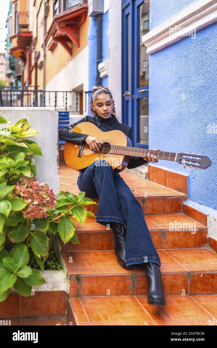 Vertikales Foto einer Afrofrau, die Gitarre spielt, auf einer Treppe vor einem bunten Haus sitzt Stockfoto