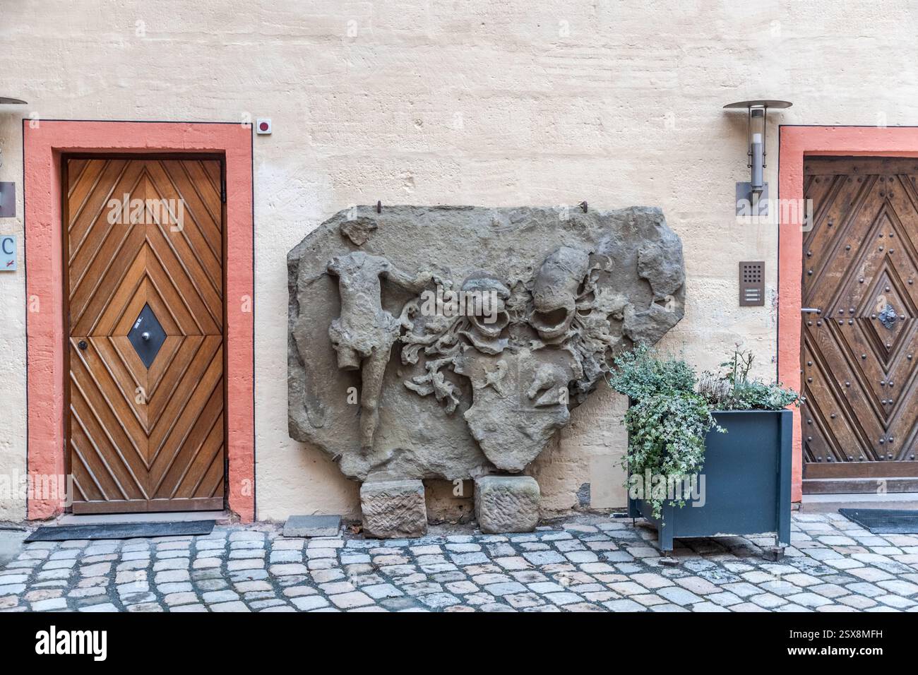 Ein verwittertes Steinrelief mit kaum erkennbaren Figuren hängt zwischen zwei geschnitzten Holztüren im Innenhof der Kaiserpfalz in Forchheim. Daneben steht ein Pflanzgefäß mit Grünpflanzen. Der Fürstbischof Lambert von Brunn aus Bamberg ließ den Komplex im späten 14. Jahrhundert errichten. Heute beherbergt das Gebäude das Pfalzmuseum. Forchheim, Bayern, Deutschland. Stockfoto