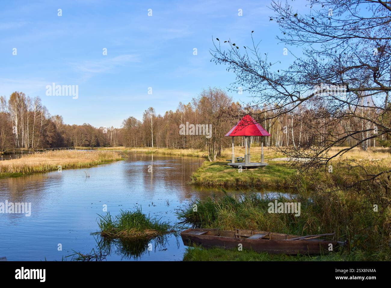Eine ruhige Seeszene mit einem Pavillon mit rotem Dach, umgeben von Herbstbäumen und einem Holzboot am grasbewachsenen Ufer Stockfoto