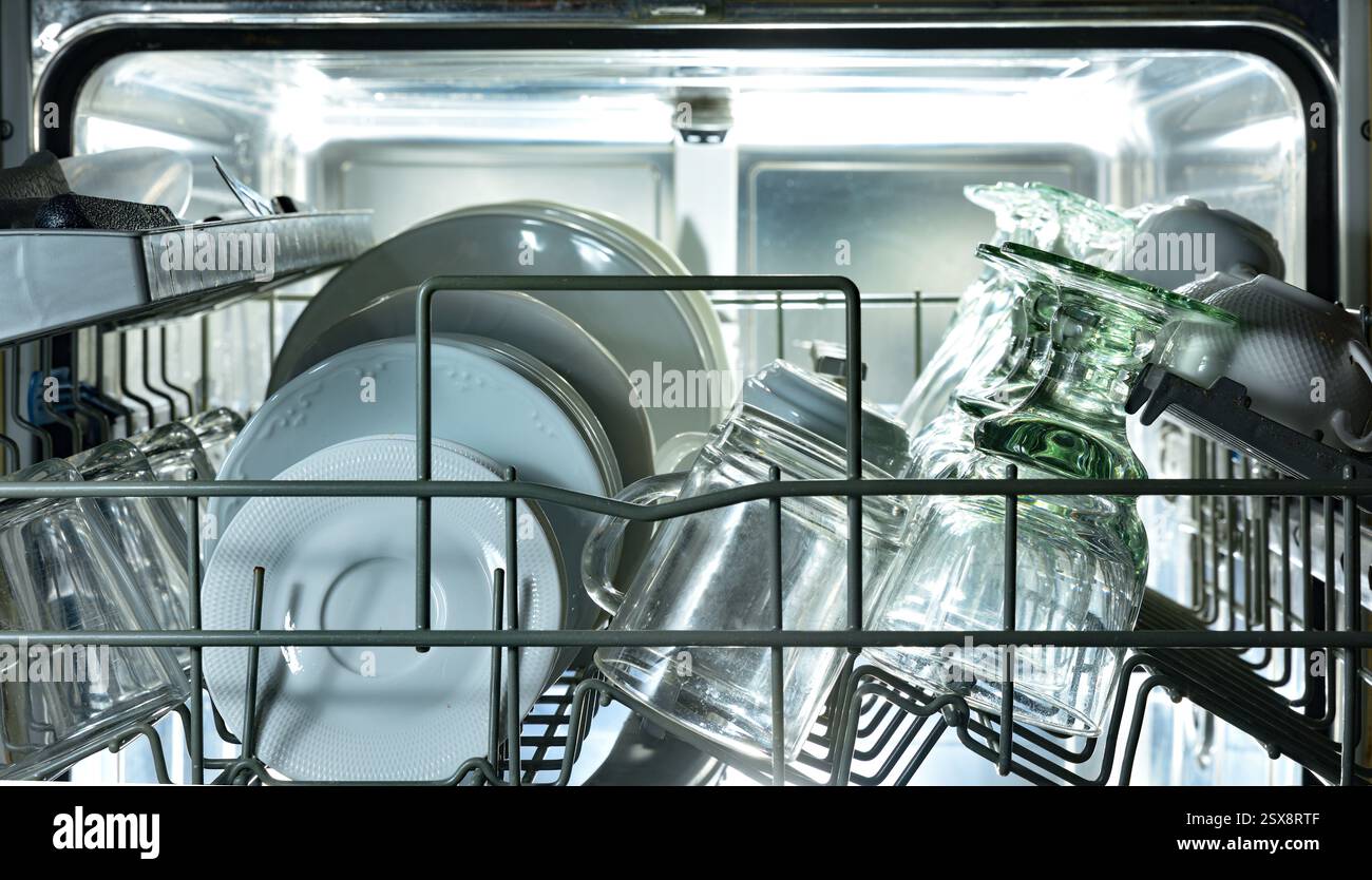 Detail des oberen Geschirrspülmaschinenregals mit Metallplatten, gefüllt mit frisch gewaschenen Tellern, Tassen und Gläsern mit Hintergrundbeleuchtung. Vorderansicht. Stockfoto