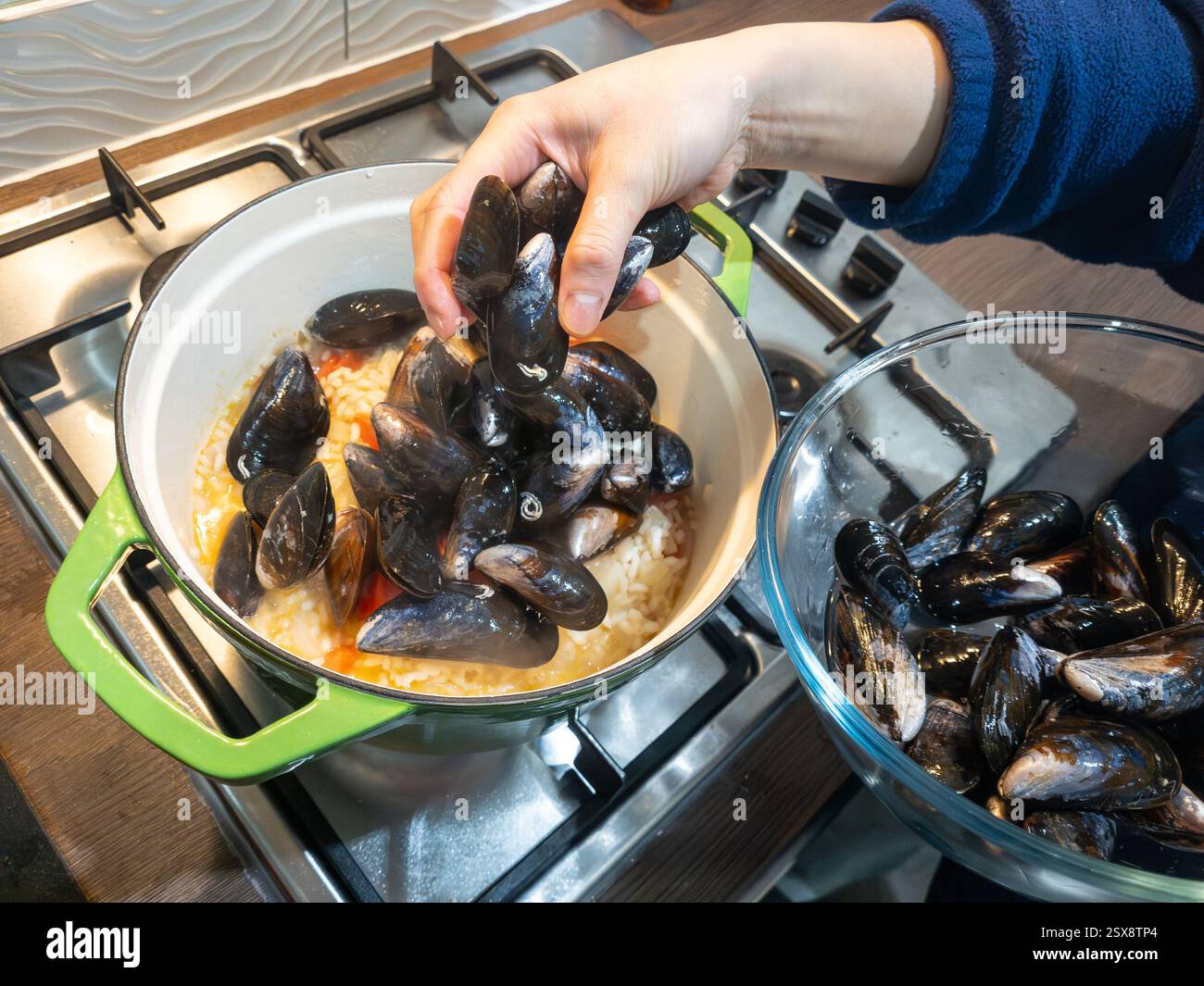 Eine Frau, die Muscheln in ihre Muscheln in Risottoreis in einem Topf auf einem Gasherd legt, um ein Risottogericht für Meeresfrüchte herzustellen, Großbritannien. Konzept: Hausmannskost - Smartphone-aufgenommenes Stockfoto