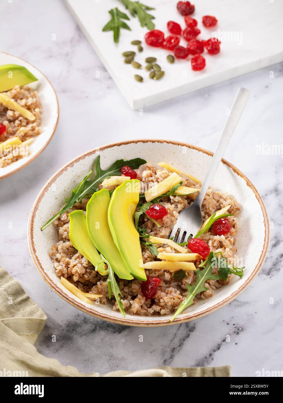 Teller mit Brei aus gemahlenem Buchweizengrieß mit Avocado, Äpfeln, getrockneten Kirschen und Salat auf dem Tisch Stockfoto