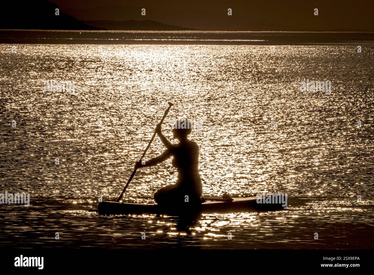 Silhouette einer Frau, die bei Sonnenuntergang paddelt und in einem ruhigen Moment des Gleichgewichts und der Freiheit durch glitzerndes goldenes Wasser gleitet Stockfoto