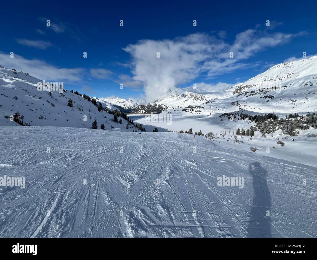 Eine atemberaubende alpine Winterlandschaft mit Skipisten, schneebedeckten Bergen und einem gefrorenen See unter einem strahlend blauen Himmel, die das Wesen einfangen - Smartphone-aufgenommenes Stockfoto