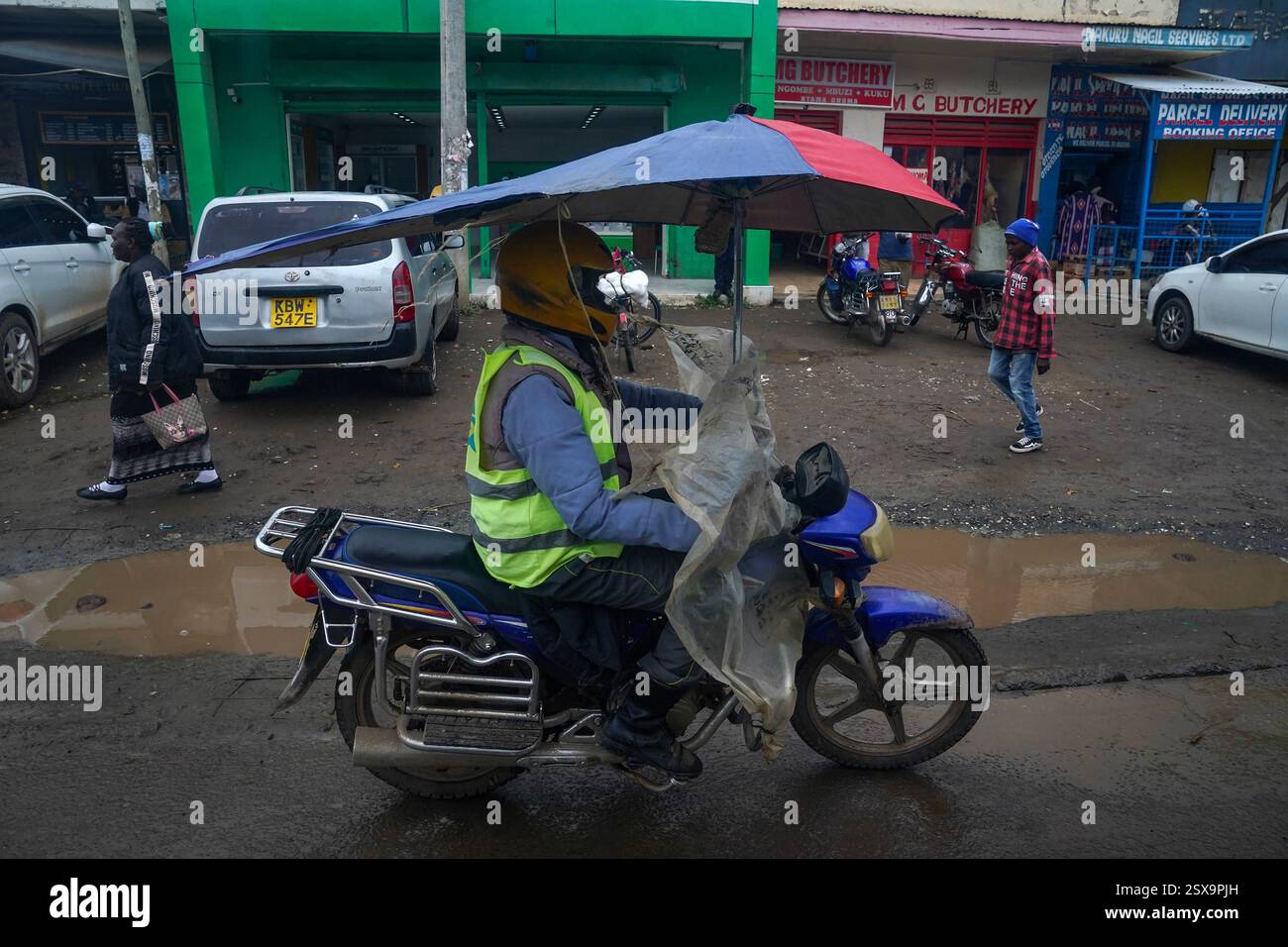 Das tägliche Leben in Gilgil, Kenia am 30. Januar 2025 fährt Ein Mann unter einem Regenschirm auf einem Motorrad in Gilgil, Kenia. Gilgil ist eine kleine, aber lebendige Stadt im Nakuru County, Kenia, am Rande des Great Rift Valley. Gilgil ist traditionell für seine landwirtschaftlichen Wurzeln bekannt und hat sich zunehmend zu einem Drehkreuz für Kleinunternehmen entwickelt, was die umfassenderen wirtschaftlichen Veränderungen in Kenia widerspiegelt. Lokale Unternehmer nutzen die robuste Telekommunikationsinfrastruktur der Nationen, die für Innovationen wie mobile Gelddienste bekannt ist, um sich mit breiteren Märkten zu verbinden und den Betrieb zu optimieren. Diese digitale Trans Stockfoto