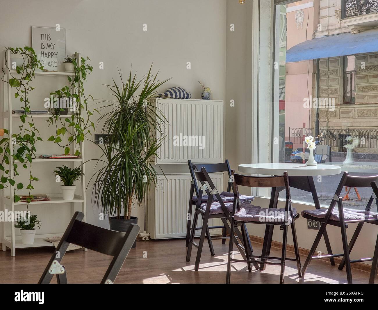 Helles, sonniges Interieur mit weißen Möbeln, Holzfaltstühlen und grünen Pflanzen; an der Wand Schild mit dem Text „This is my Happy Place“; Fenster Stockfoto