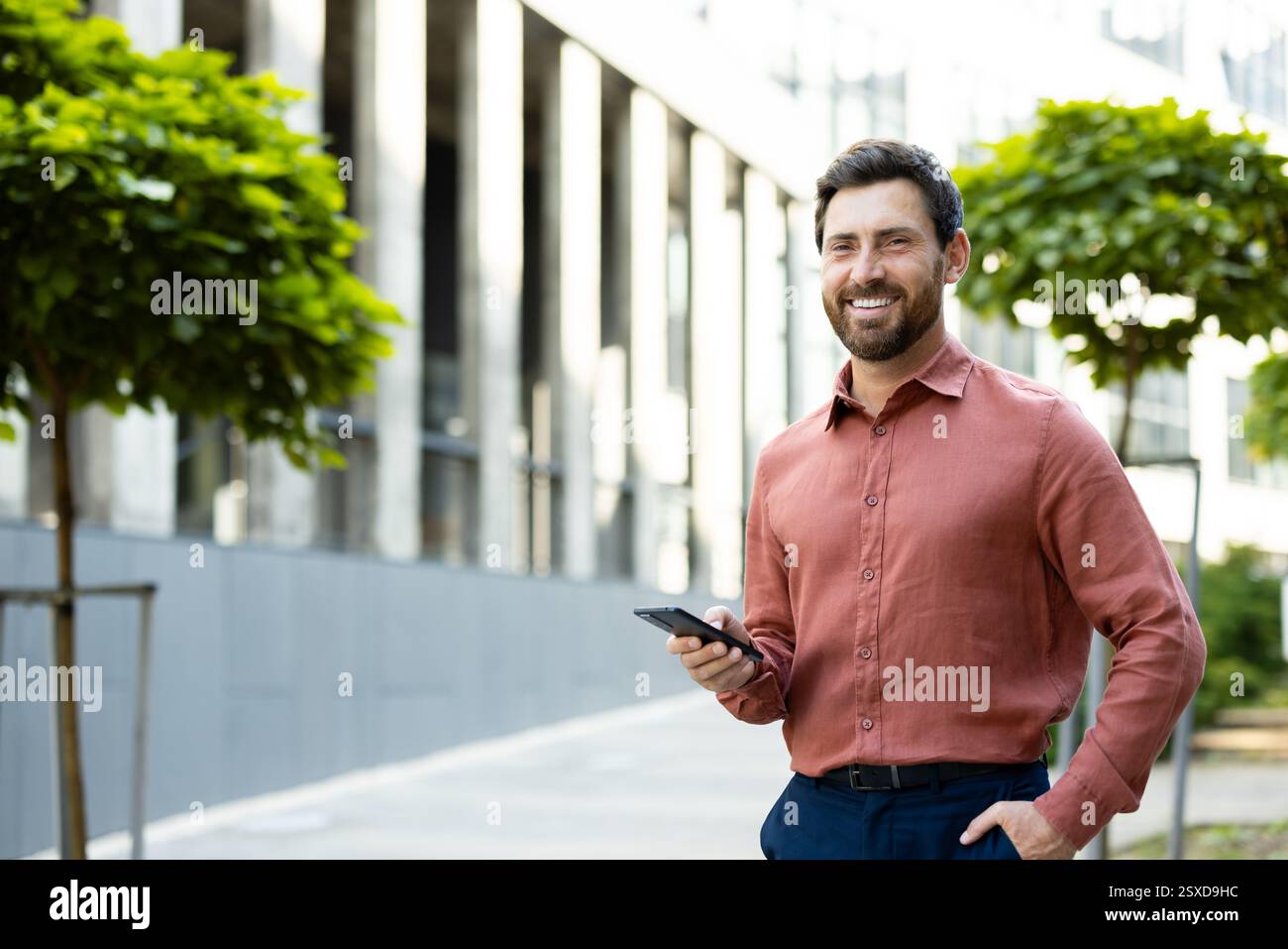 Porträt eines erfolgreichen und freudigen Geschäftsmannes. Ein Mann mit einem Telefon in der Hand lächelt und schaut in die Kamera, während er aus einem Bürogebäude geht. Stockfoto
