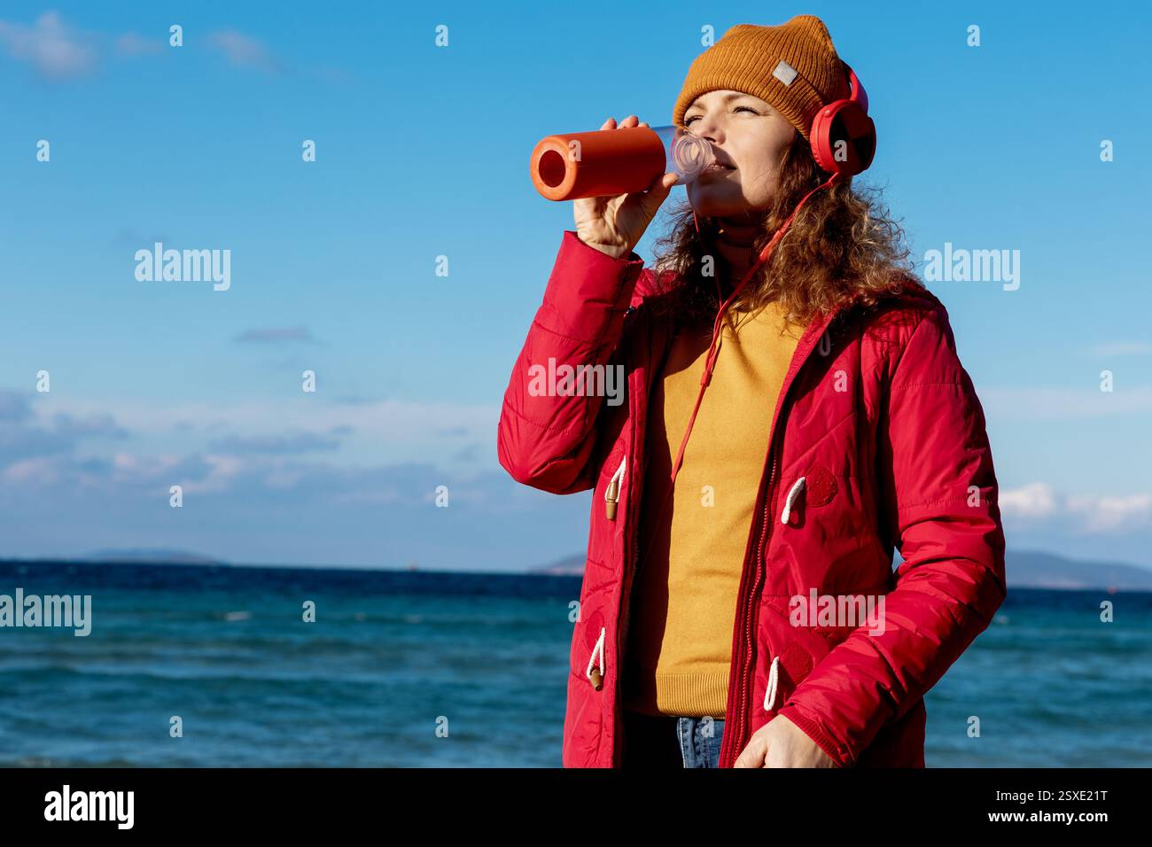 Junge Frau mit Kopfhörern, die bei kaltem Wetter Wasser am Meer trinken Stockfoto