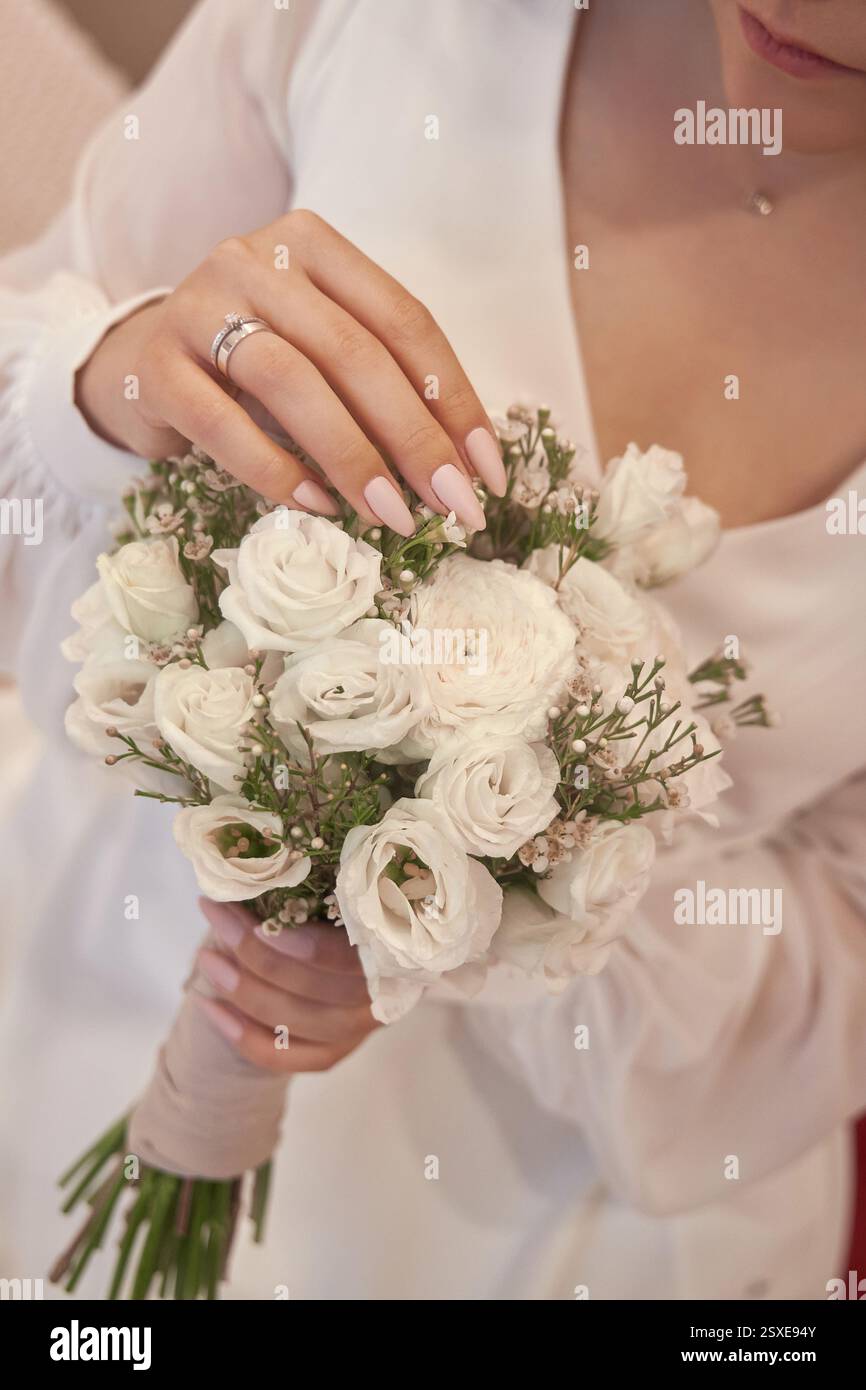 Braut arrangiert liebevoll ihren atemberaubenden weißen Blumenstrauß, gefüllt mit zarten Blumen und Grün, Momente vor ihrer Hochzeitszeremonie in einem eleganten B Stockfoto