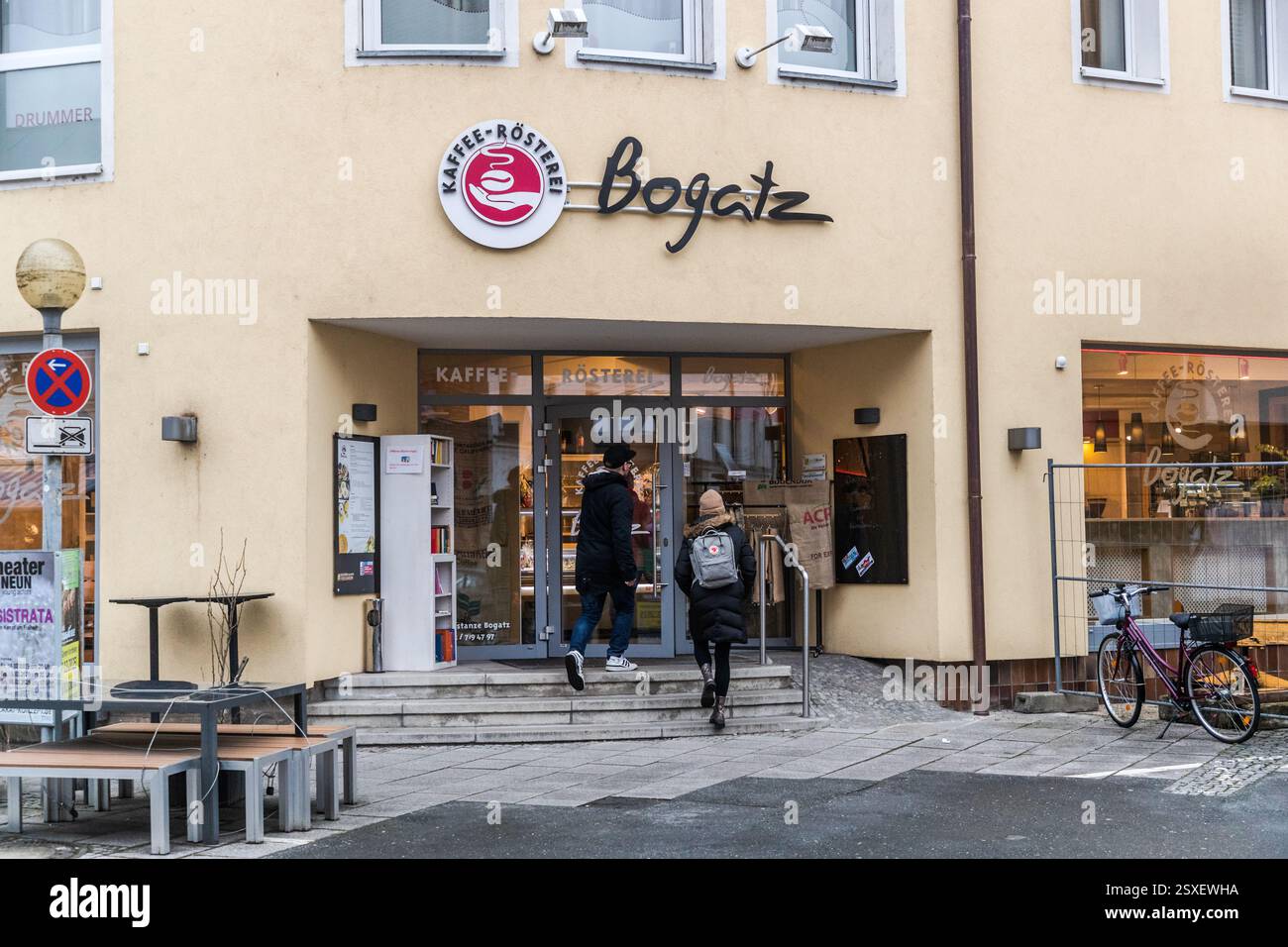 Die Bogatz-Kaffeerösterei in der Altstadt von Forchheim begrüßt zwei Fußgänger unter ihrem runden roten Logo über dem Eingang. Bänke und ein kleiner Tisch stehen vor der cremefarbenen Fassade, rechts lehnt ein Fahrrad an der Wand. Die Rösterei röstet Kaffeebohnen aus Ländern wie Brasilien, Guatemala, Indien und Äthiopien in einer traditionellen Trommelröstmaschine. Hornschuchallee, Forchheim, Bayern, Deutschland. Stockfoto