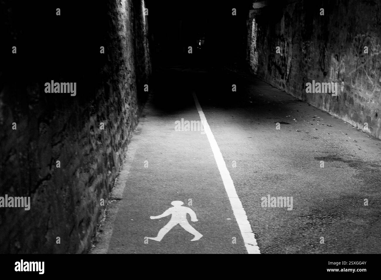 Ein Fußgängerweg, der mit einer weißen Linie und einem Symbol für eine Wanderfigur markiert ist, führt in eine dunkle und enge Gasse, die auf eine ausgewiesene Route hinweist. Stockfoto