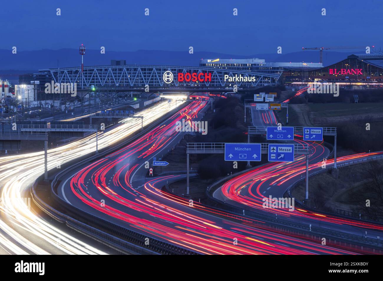 Autobahn A8 am Flughafen Stuttgart am Abend. Starker Verkehr, leichte Fahrspuren. Moderne Architektur der Messe Stuttgart mit Parkhaus Stockfoto