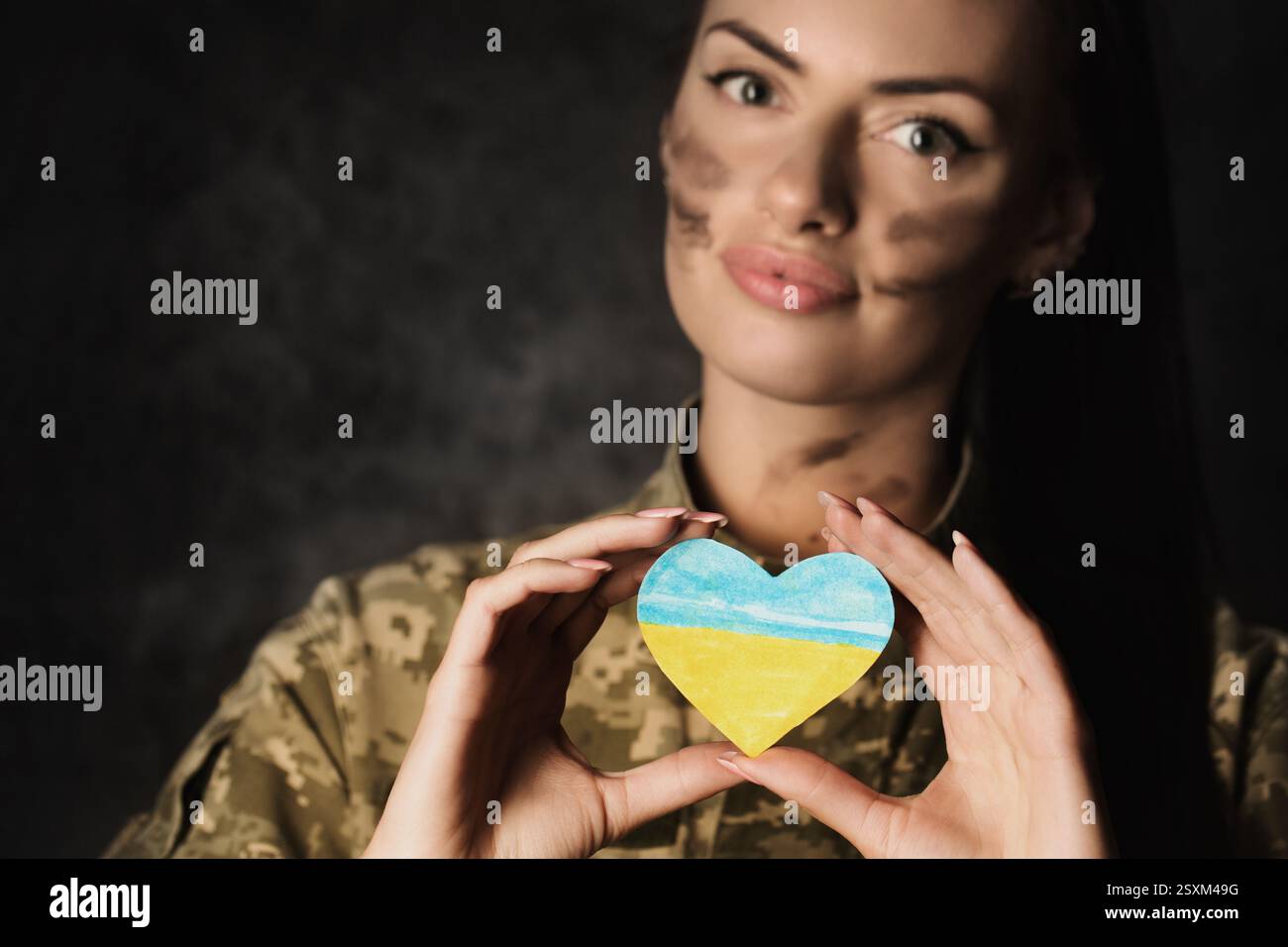 Schöne junge Militärfrau in Militäruniform mit Tarnschminke im Gesicht hält eine herzförmige ukrainische Flagge in den Händen. Studiofoto o Stockfoto