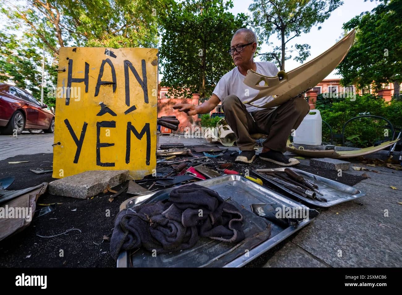 Ein Mann, der auf dem Bürgersteig sitzt, repariert kleine Autodefekte nach Unfällen neben einer Straße im Zentrum von Hanoi. Stockfoto