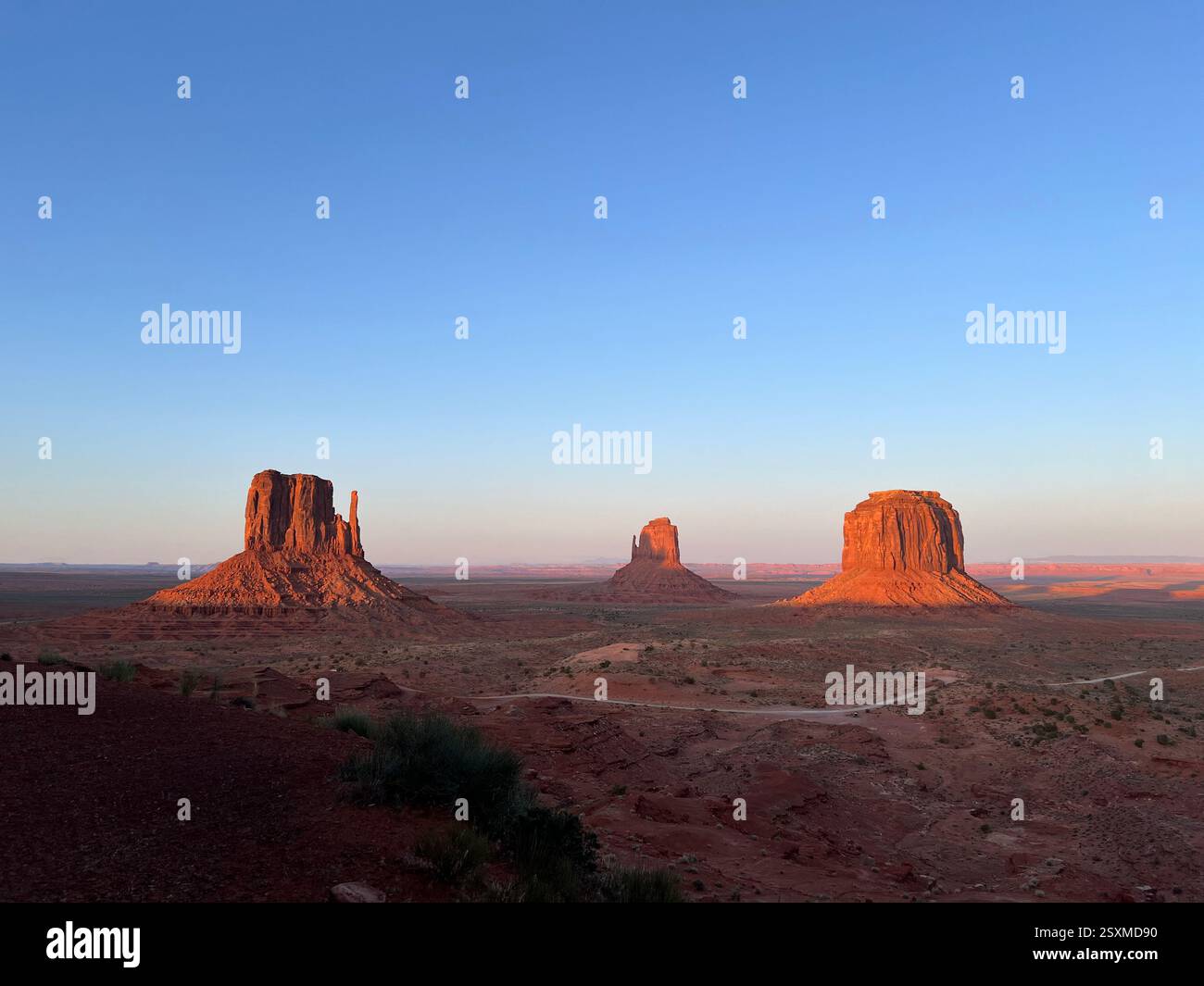 Der Sonnenuntergang wirft ein feuriges Leuchten auf die drei Monolithen des Monument Valley, während roter Sand und der Valley Scenic Drive im Schatten unter einem klaren Himmel ruhen. - Smartphone-aufgenommenes Stockfoto