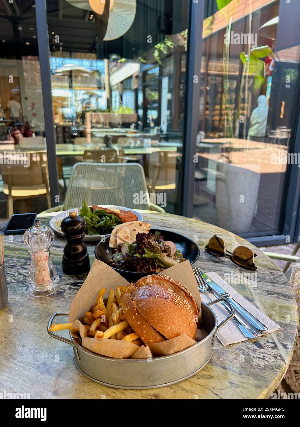 Das Bild zeigt einen Restauranttisch im Freien mit drei Gerichten: Einen Burger mit Pommes Frites in einem Metalltablett, einen Falafelteller mit Salat und Fladenbrot und einen Sc Stockfoto