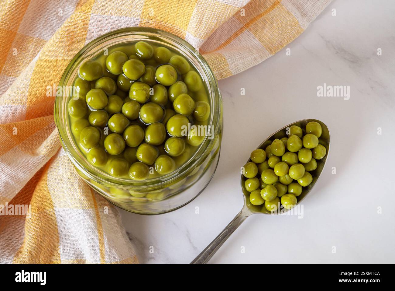 Voller Becher mit grünen Erbsen in Dosen auf Marmorplatte. Nahaufnahme marinierter Erbsen in einem Glasgefäß und einem Metalllöffel. Snack, Salatzutat, Gemüse Stockfoto