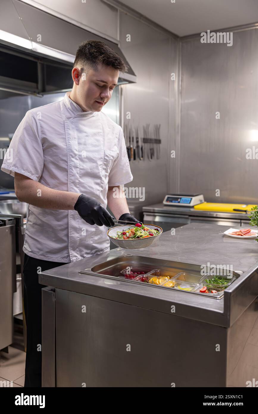 Junger Koch, der frischen Salat in der Restaurantküche mit verschiedenen Zutaten zubereitet Stockfoto
