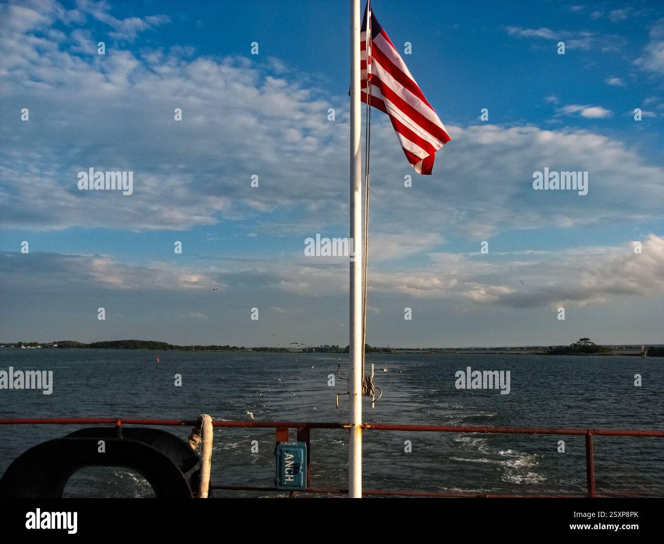 Eine Fahne flattert im Wind von einem weißen Fahnenmast auf dem Heck eines Tankers. Das Schiff bewegt sich und hinterlässt eine schaumige Spur. Der Himmel ist eine Mischung aus Stockfoto