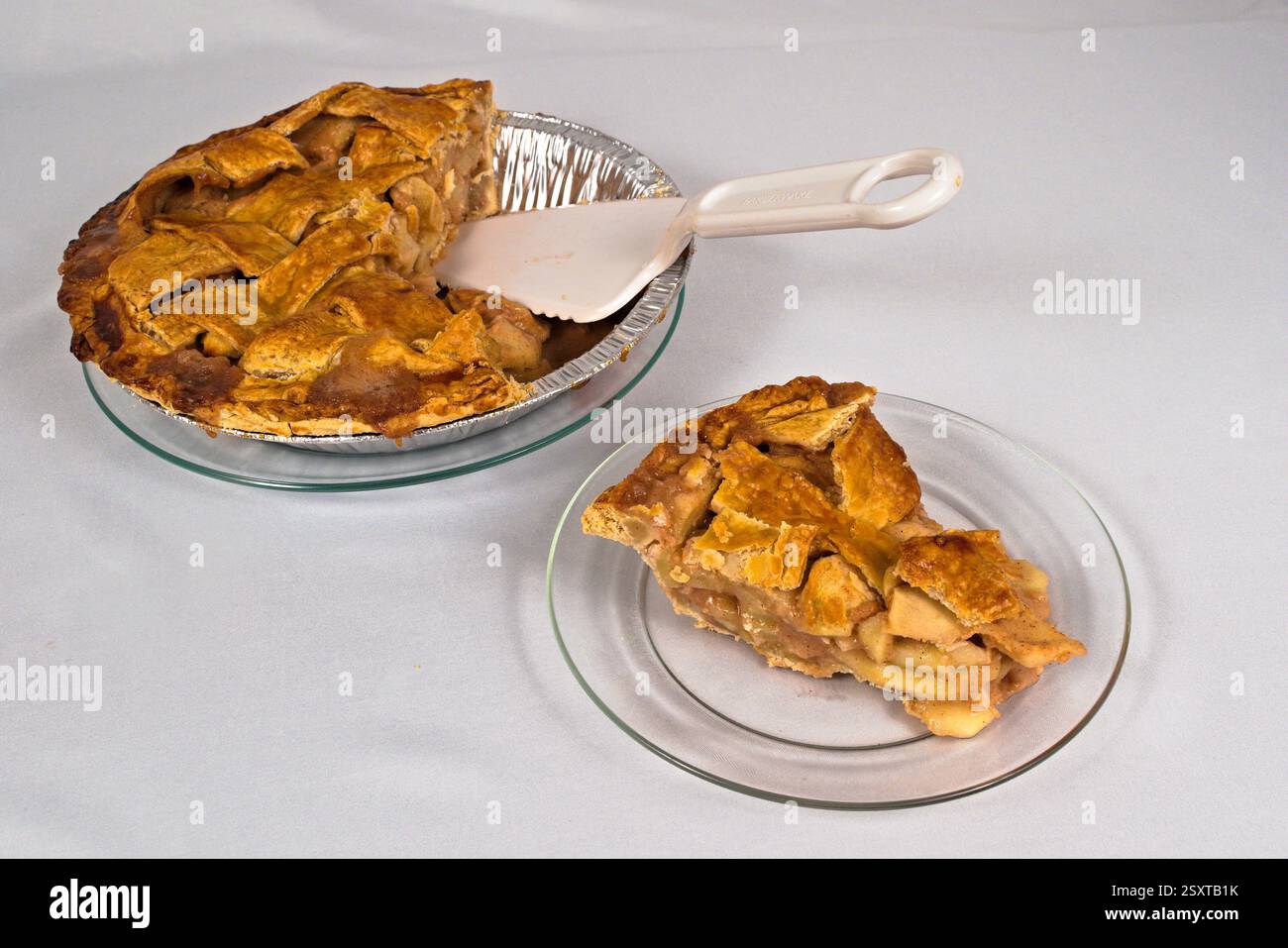 Ein köstlicher hausgemachter Apfelkuchen mit einer Scheibe auf einer Glasplatte neben einem Tortenschneider. Weißer Hintergrund. Stockfoto