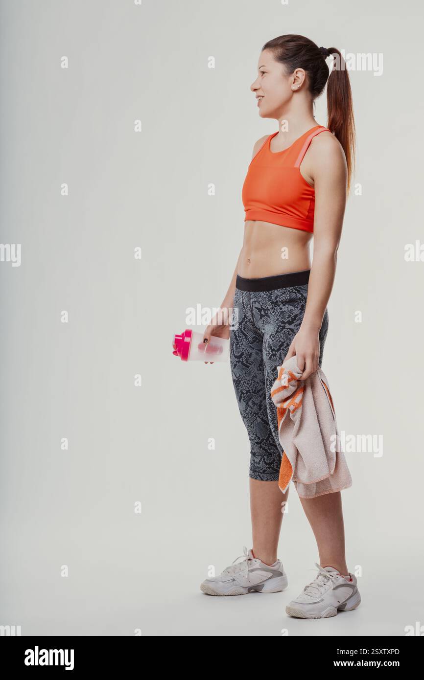 Junge sportliche Frau, die im Profil steht, eine Wasserflasche und ein Handtuch hält und nach einem belebenden Workout erfrischt und Energie ausstrahlt Stockfoto