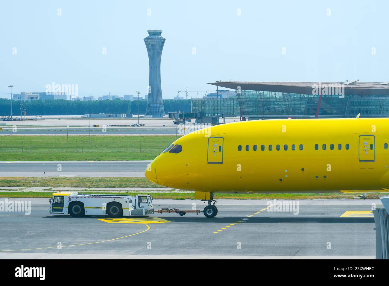 Ein gelbes Flugzeug wird von einem Fahrzeug auf der Landebahn eines Flughafens abgeschleppt. Im Hintergrund sind ein modernes Terminal und ein Steuerturm unter einem sichtbar Stockfoto