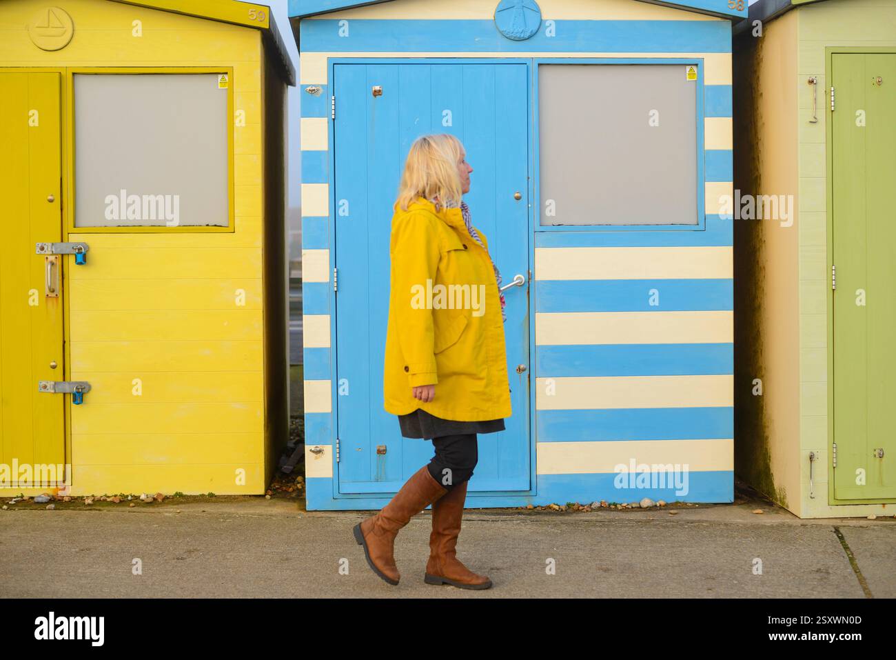 Frau im gelben Mantel, die an einer blauen Strandhütte vorbeiläuft Stockfoto