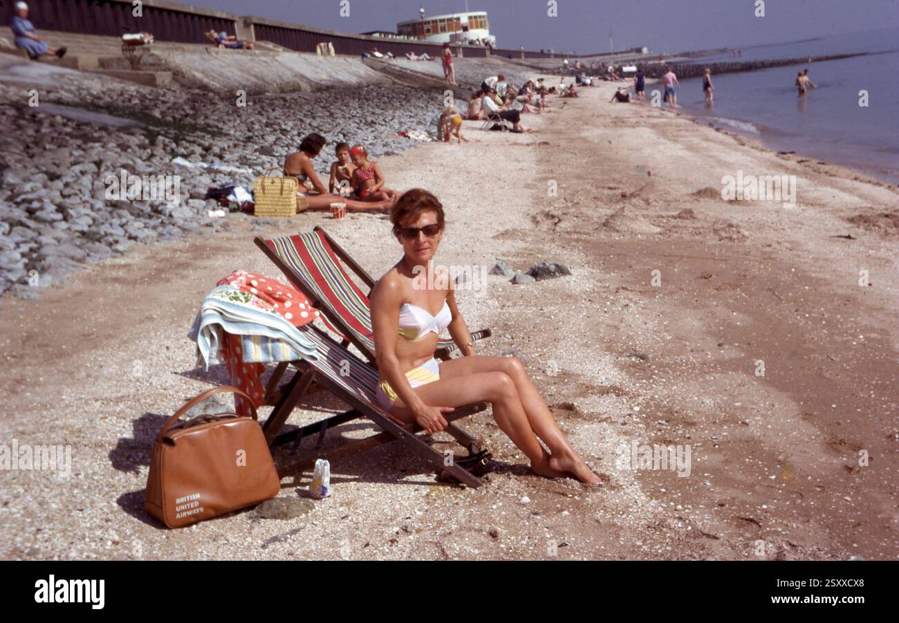 Frau in einem weißen Bikini auf einem Liegestuhl am Strand Nest zu einer British Unted Airways Tasche, Canvey Island, 1967 Foto vom Henshaw Archive Stockfoto