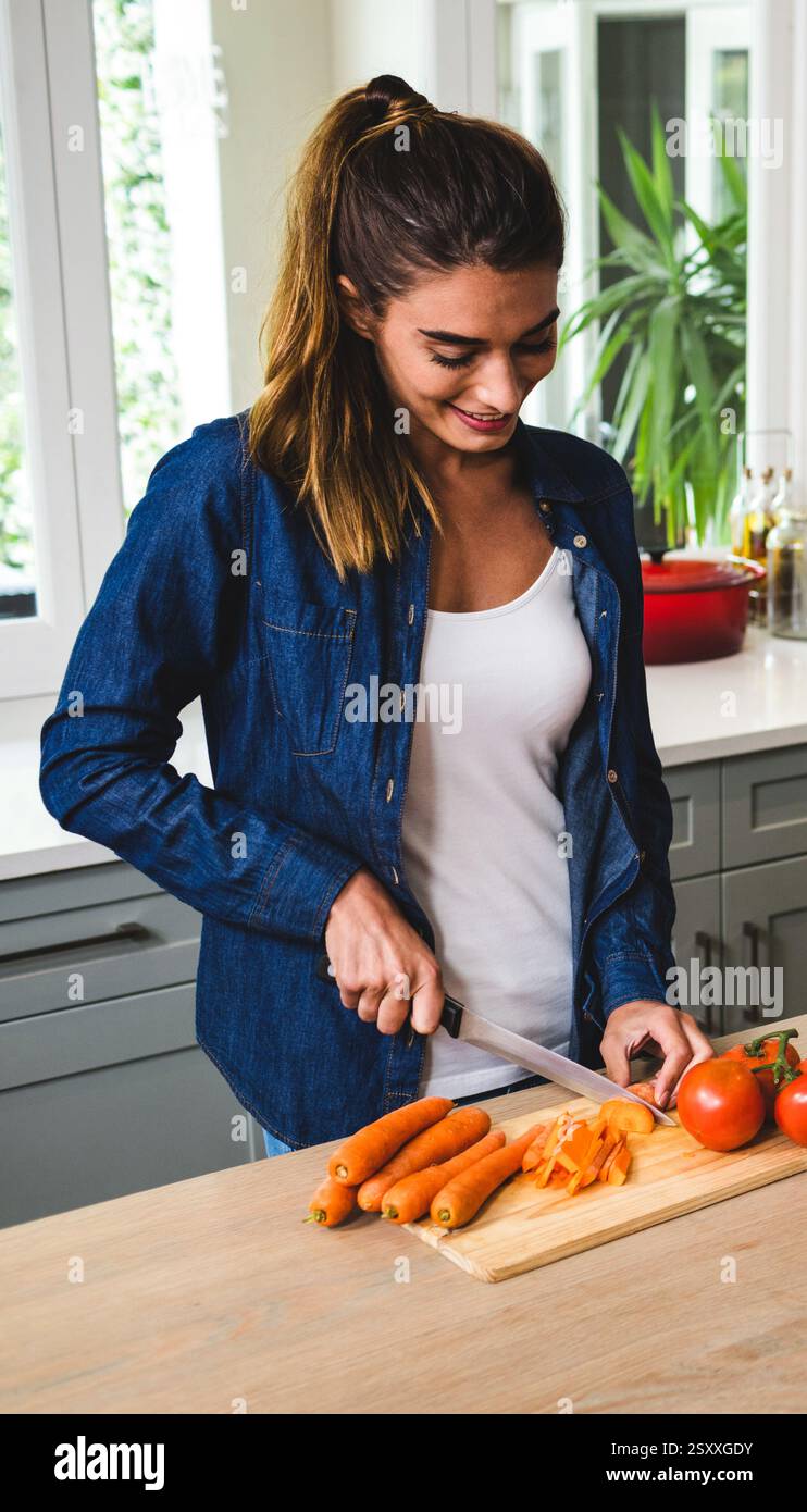 Lächelnde Frau, die Karotten in der Küche hackt und frische Zutaten zum Kochen zubereitete Stockfoto
