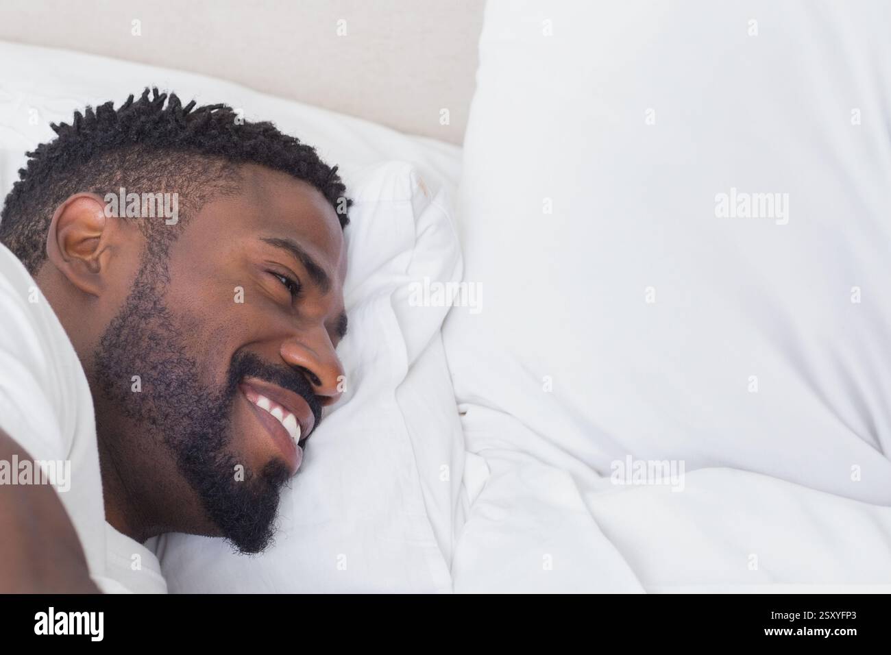 Afroamerikaner lächelnd, während er sich bequem auf weißer Bettwäsche und Kopierraum entspannte Stockfoto
