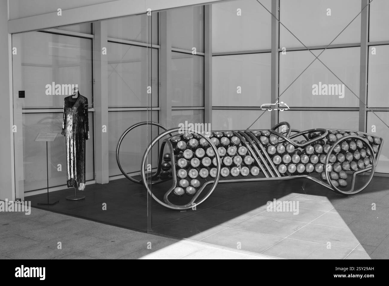 Málaga, Spanien - 28. Juni 2024: Kleid und eine Autoskulptur aus Edelstahl und Scheinwerfern im Automobil- und Modemuseum Malaga. Stockfoto