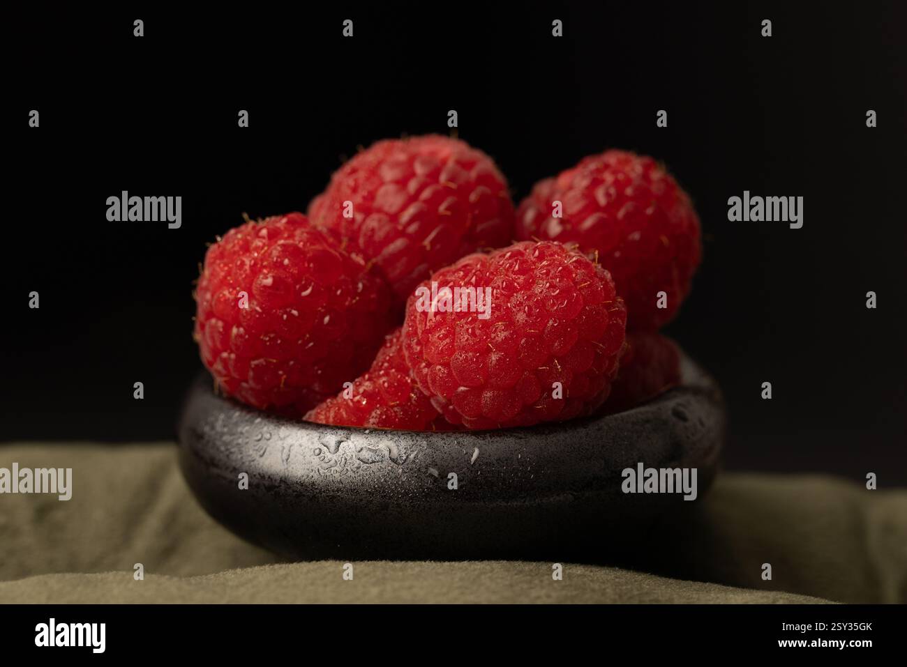 Frische Himbeeren auf einer winzigen, schwarz glänzenden Schale und auf einer grünen Tischdecke. Dramatische Studiobeleuchtung. Stockfoto