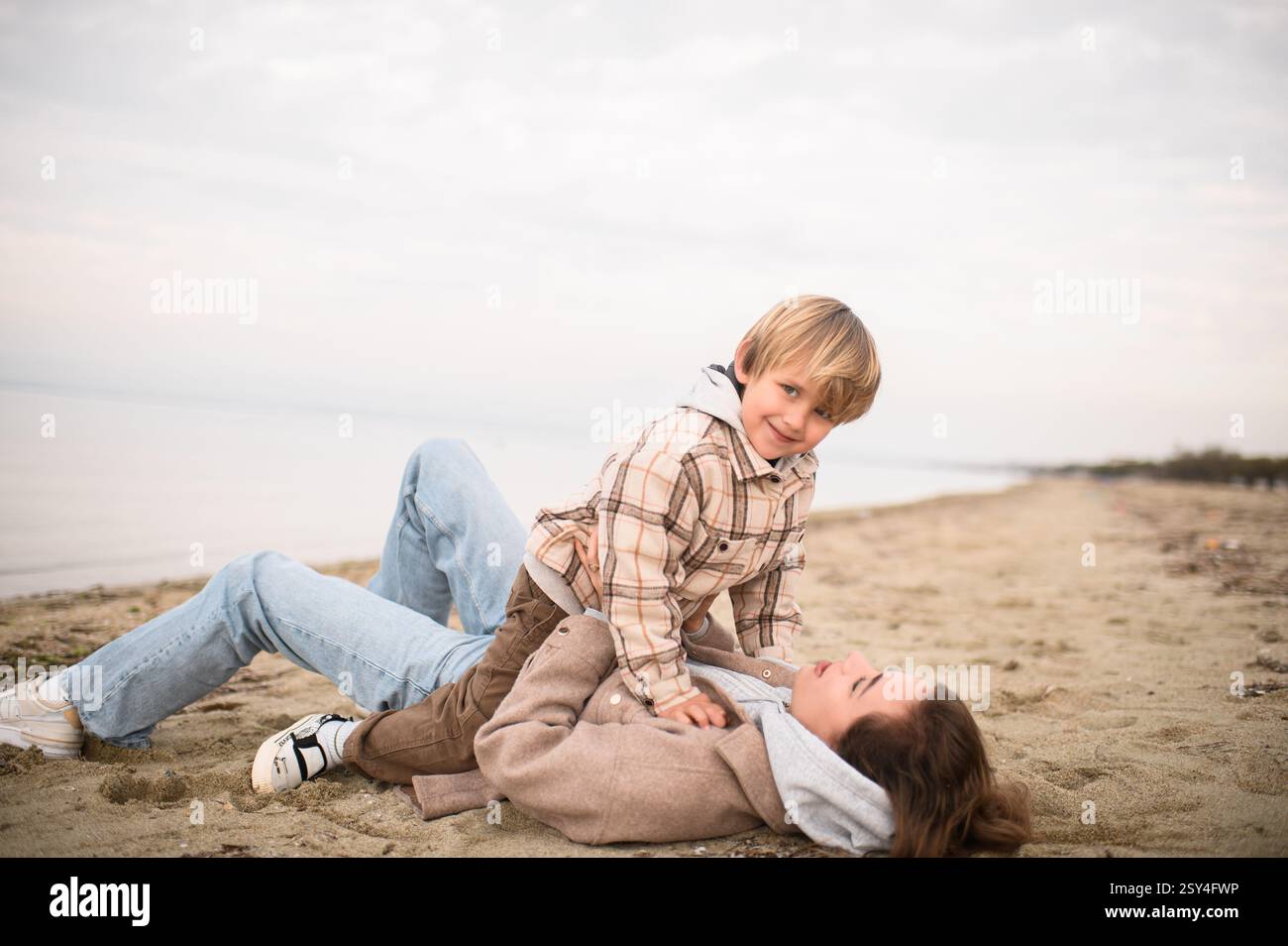Die Mutter liegt auf dem Sand, hebt ihren Sohn über sie, während er seine Arme ausstreckt und so tut, als würde er fliegen, beide lachen in der salzigen Meeresbrise. Stockfoto