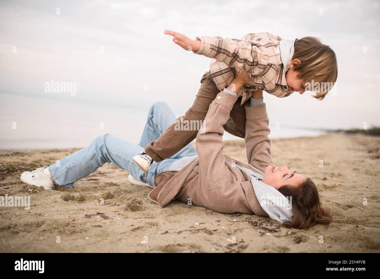 Die Mutter liegt auf dem Sand, hebt ihren Sohn über sie, während er seine Arme ausstreckt und so tut, als würde er fliegen, beide lachen in der salzigen Meeresbrise. Stockfoto