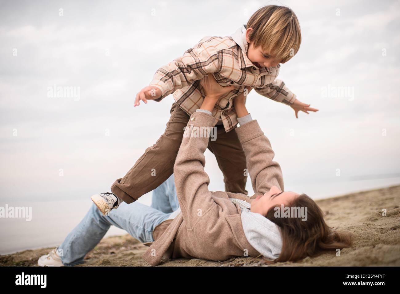 Die Mutter liegt auf dem Sand, hebt ihren Sohn über sie, während er seine Arme ausstreckt und so tut, als würde er fliegen, beide lachen in der salzigen Meeresbrise. Stockfoto
