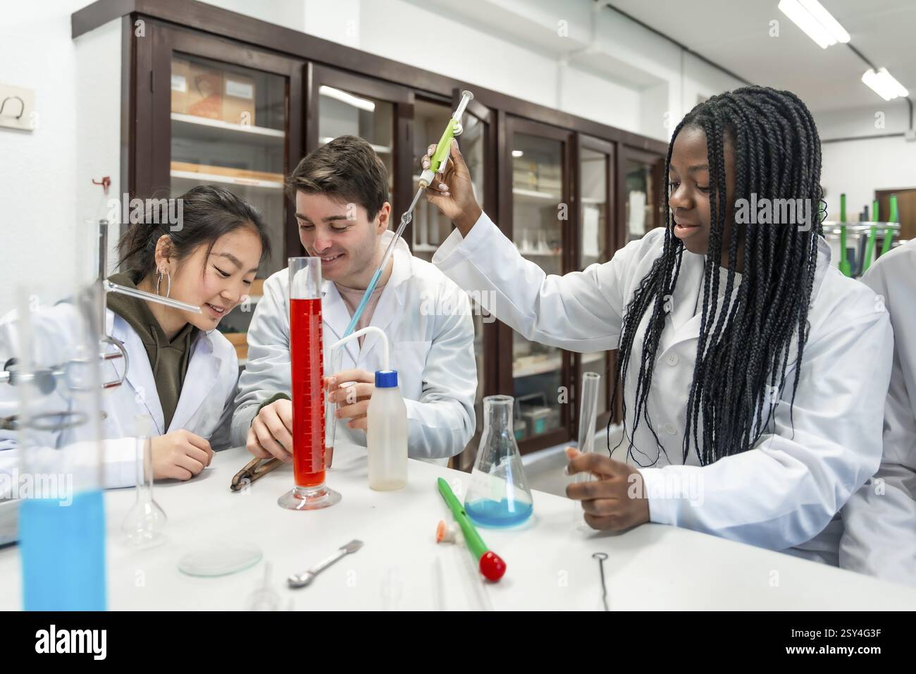 Multiethnische Universitätsstudenten in Labormänteln, die ein Chemieexperiment mit bunten Flüssigkeiten und Laborgeräten durchführen Stockfoto