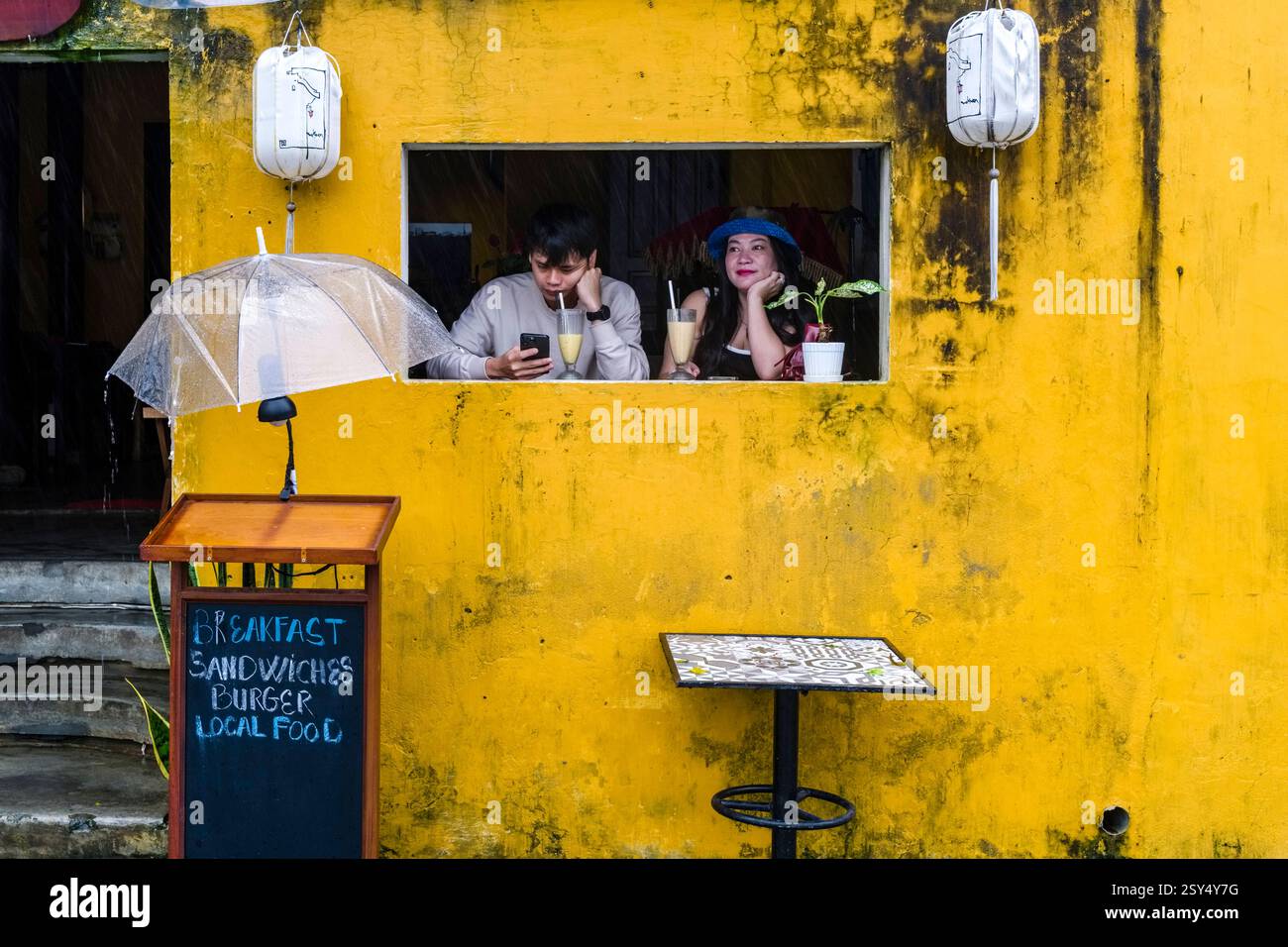 Zwei Touristen mit Getränken und Mobiltelefonen sehen aus einem gelben Haus in der Altstadt von Hoi an, Hái an einem regnerischen Tag gelangweilt aus. Provinz Hoi An Quang Nam Stockfoto