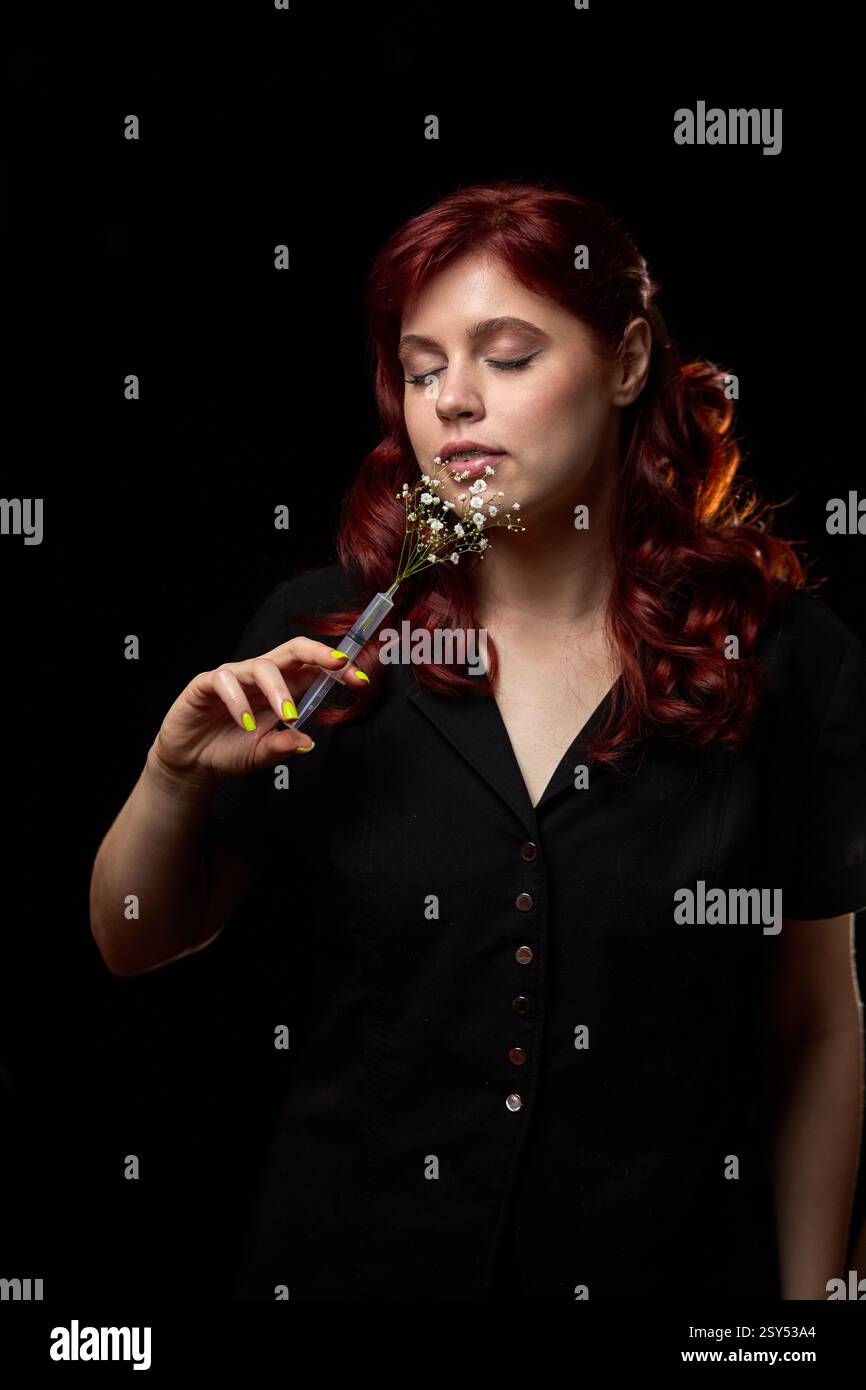 Friedliche Frau mit rotem Haar in schwarzem Kleid, während sie zarte Blumen sanft hält, während sie ihre Augen schließt und ein Gelassenheit-Konzept umschließt Stockfoto