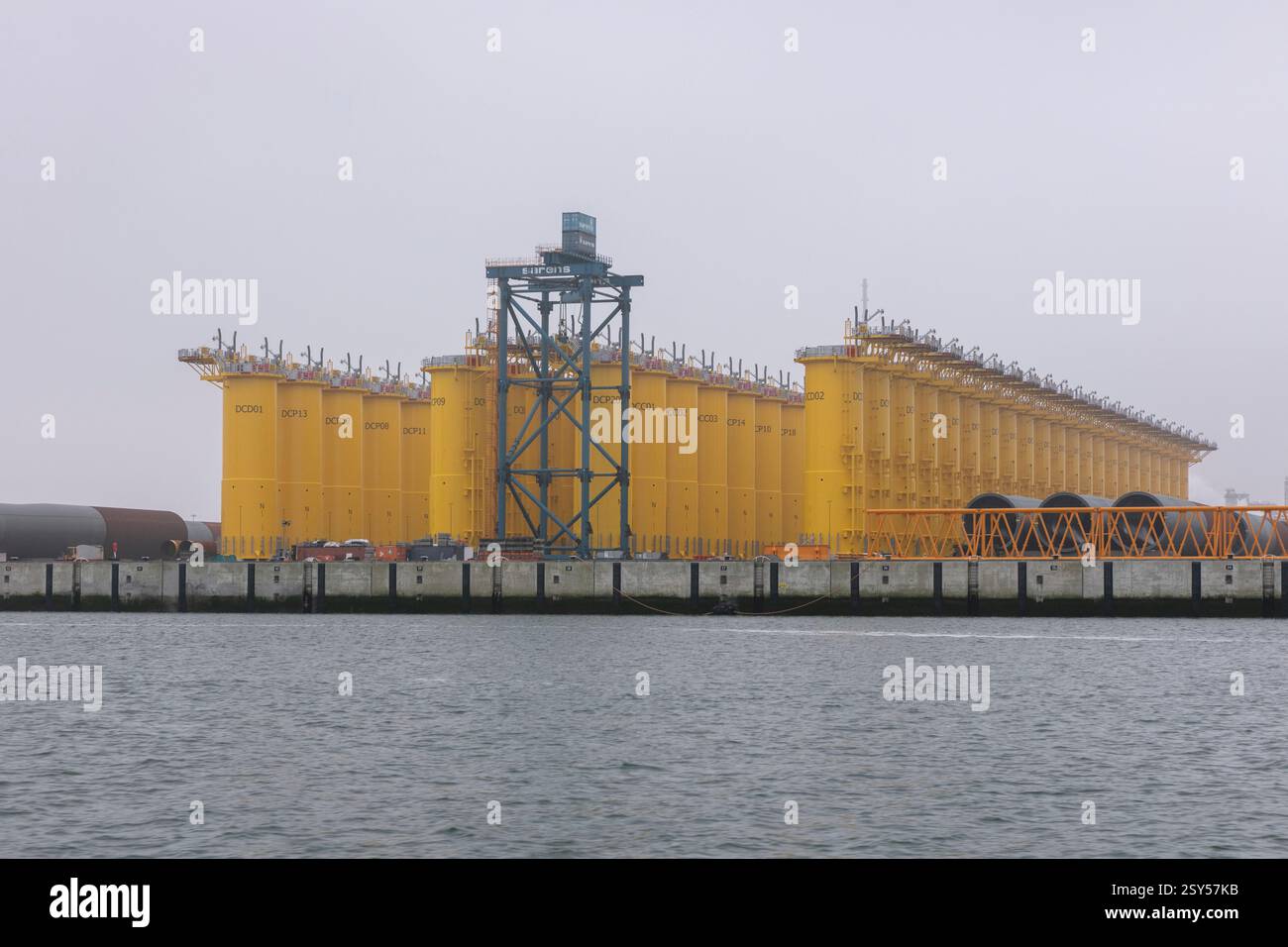Nebeliger Vormittag im Prinses Margriethaven, Rotterdam, mit einer Reihe von gelben Monopiles und Übergangsteilen, die für die Installation in einem Offshore-Windpark bereit sind. Diese Industrieszene fängt das Wesen der Entwicklung erneuerbarer Energien vor dem Hintergrund von ruhigem Wasser und gedämpftem grauem Himmel ein. Stockfoto