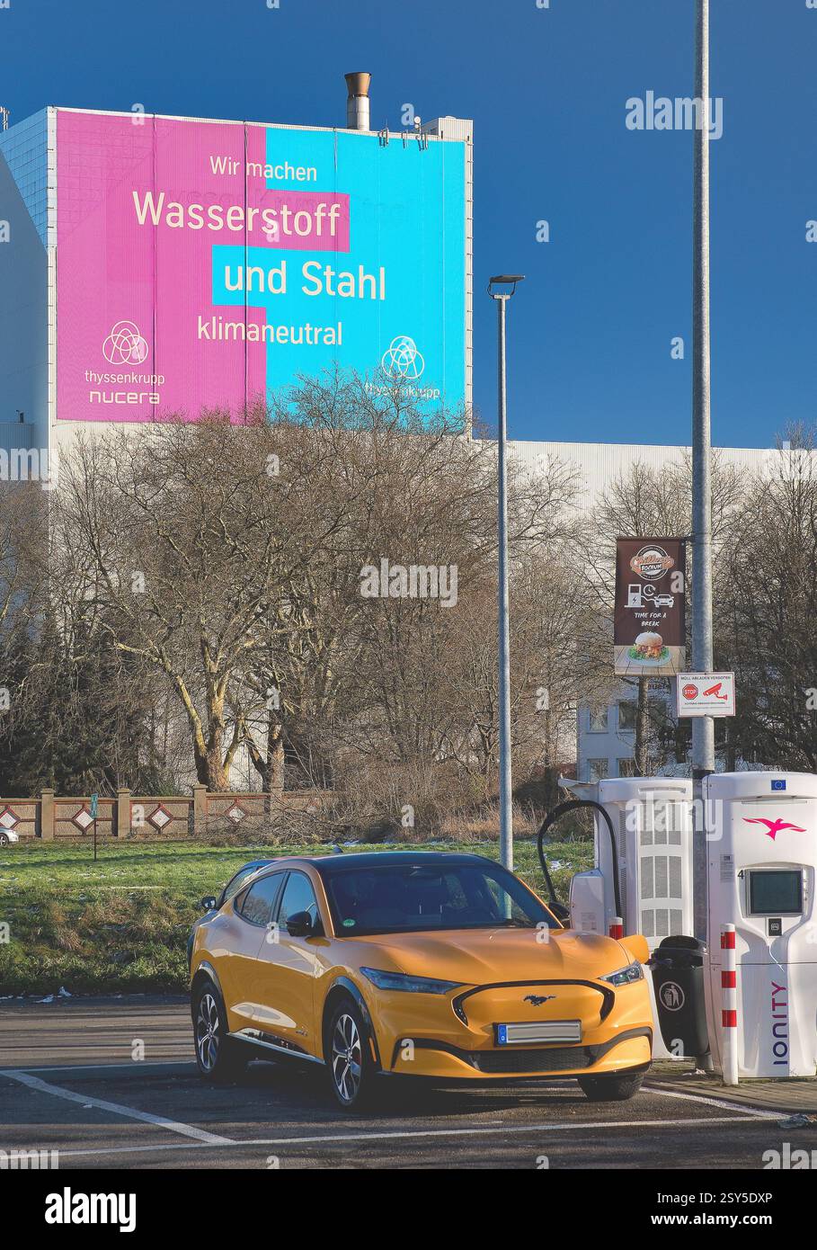 Orangefarbenes Elektroauto an der elektrischen Ladestation, großes Poster für Klimastahl im ThyssenKrupp Steel Europe Werk im Hintergrund, Deutschland Stockfoto