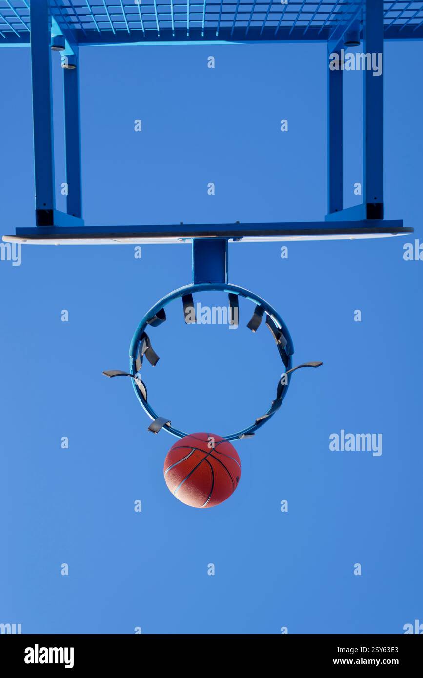 Ein Basketball läuft durch einen abgenutzten blauen Metallkorb vor einem klaren Himmel. Das Netz fehlt, zeigt eine städtische Outdoor-Sportanlage. Stockfoto