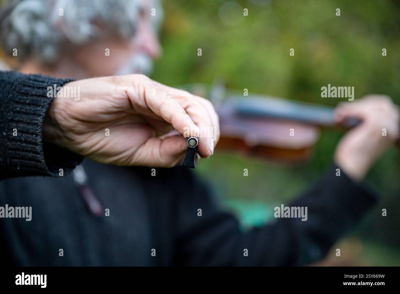Eine Nahaufnahme einer Geigerhand, die den Bogen hält, mit einem verschwommenen Hintergrund, der einen älteren Musiker zeigt, der im Freien Geige spielt. Stockfoto