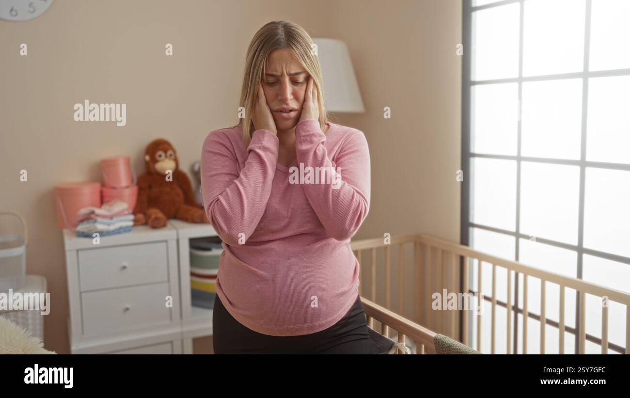Schwangere Frau, die sich gestresst fühlt, sitzt auf dem Bett in einem gemütlichen Schlafzimmer mit Wiege in der Nähe, umgeben von wichtigen Babysachen und weichem Licht, das durch die große W Stockfoto