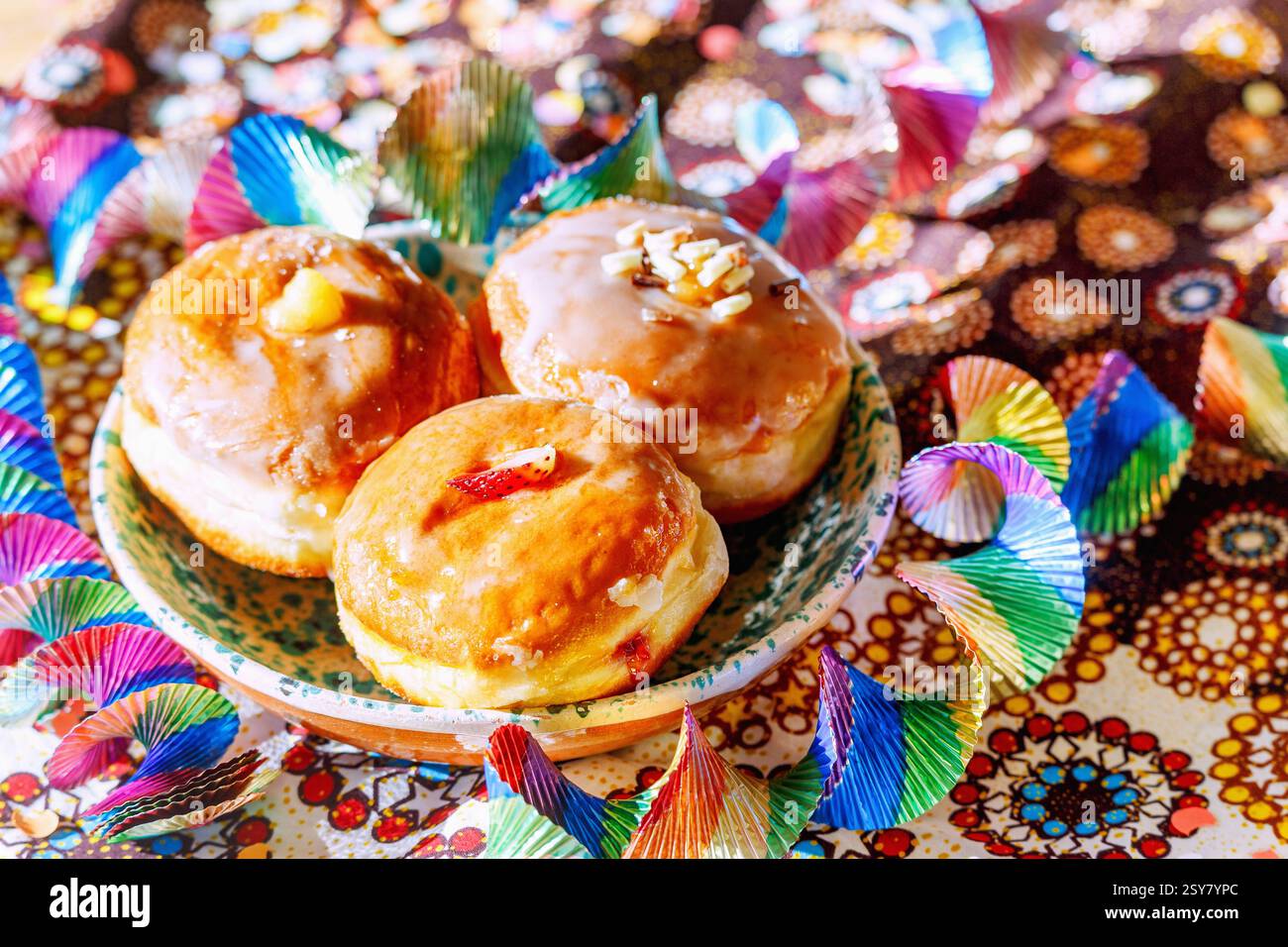 bunte gefüllte Faschingskrapfen mit Zuckerguss Erdbeer-Limes, Eierlikör, weiße Schokoladencreme, Keramikteller weiß-grün melliert, gemusterte Tischdec Stockfoto