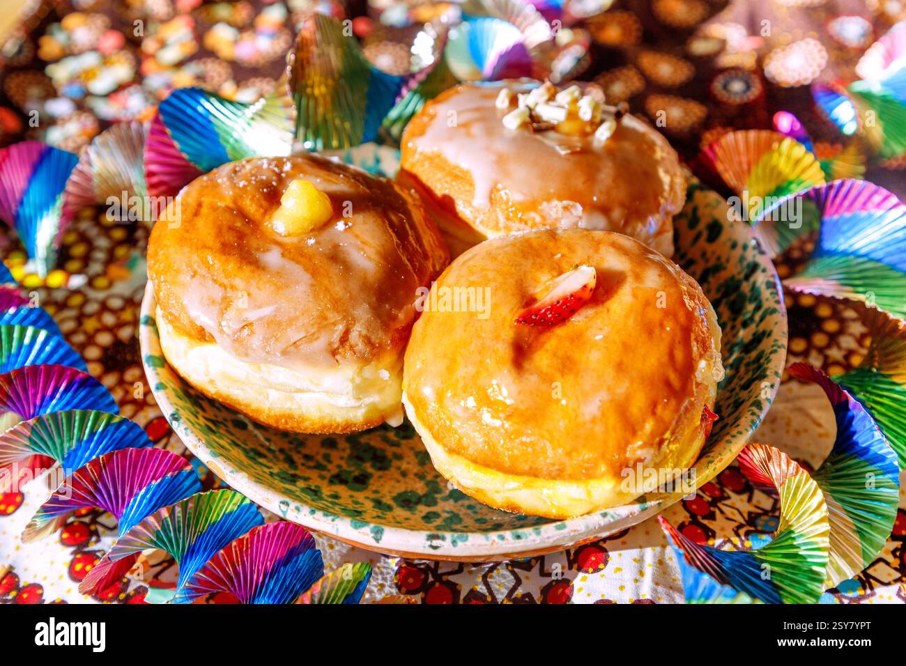 bunte gefüllte Faschingskrapfen mit Zuckerguss Erdbeer-Limes, Eierlikör, weiße Schokoladencreme, Keramikteller weiß-grün melliert, gemusterte Tischdec Stockfoto