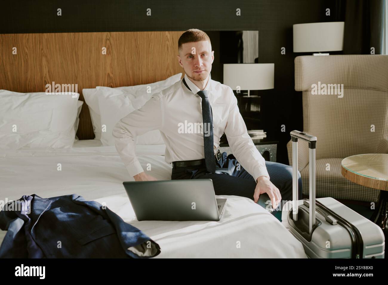 Hochwinkel-Porträt eines erfolgreichen Geschäftsmannes, der auf dem Bett sitzt, mit Laptop und Blick auf die Kamera, seinem Jack auf dem Bett und silbernem Koffer in der Nähe des Doppelbetts Stockfoto