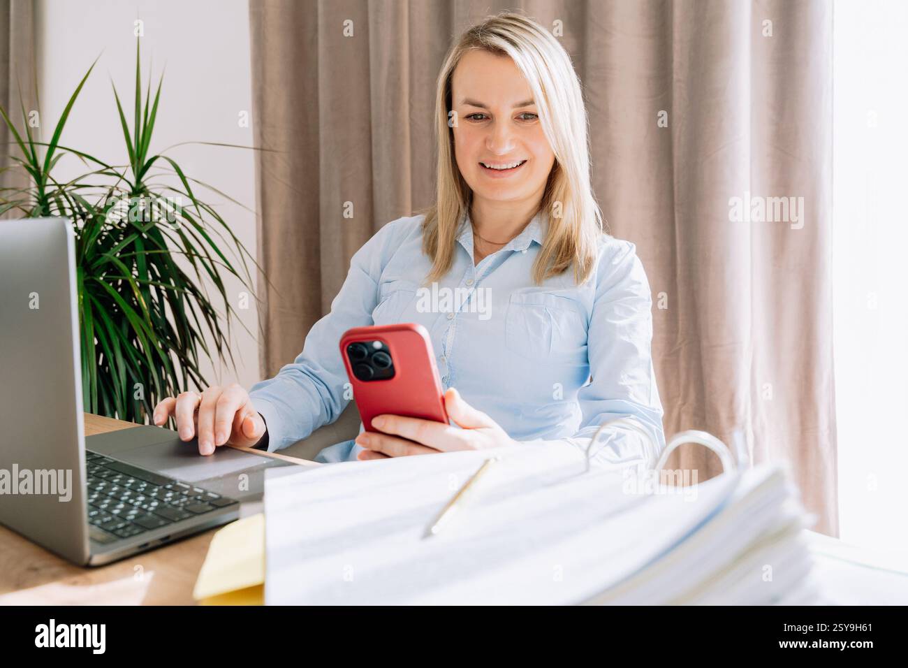 Ein lächelndes Mädchen, das ein Telefon hält, sitzt am Tisch neben dem Laptop und hinter einem Ordner mit Dokumenten, 35-37 Jahre altes europäisches Aussehen aus der Nähe Stockfoto