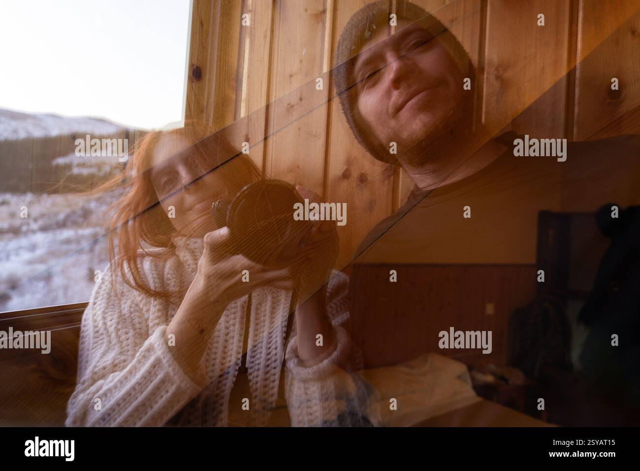 Eine gemütliche Winterszene zeigt eine rothaarige Frau mit einer Kamera, die neben einem Mann in einem Glasfenster vor einem verschneiten Hintergrund reflektiert wird Stockfoto