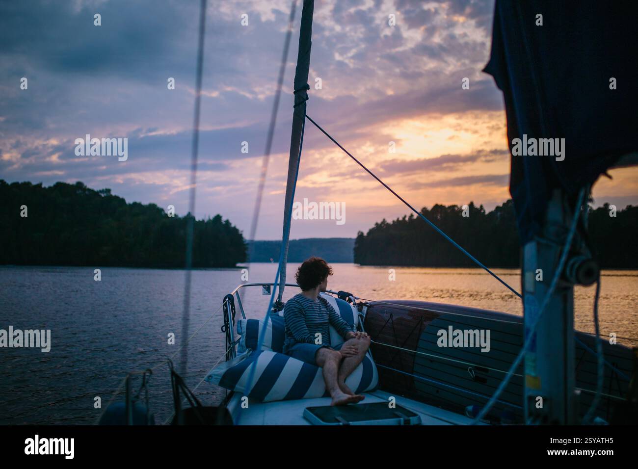 Ein ruhiger Moment auf einem Segelboot bei einem malerischen Sonnenuntergang. Ein Mann entspannt sich an Deck und genießt die Ruhe des ruhigen Wassers und des lebendigen Himmels Stockfoto