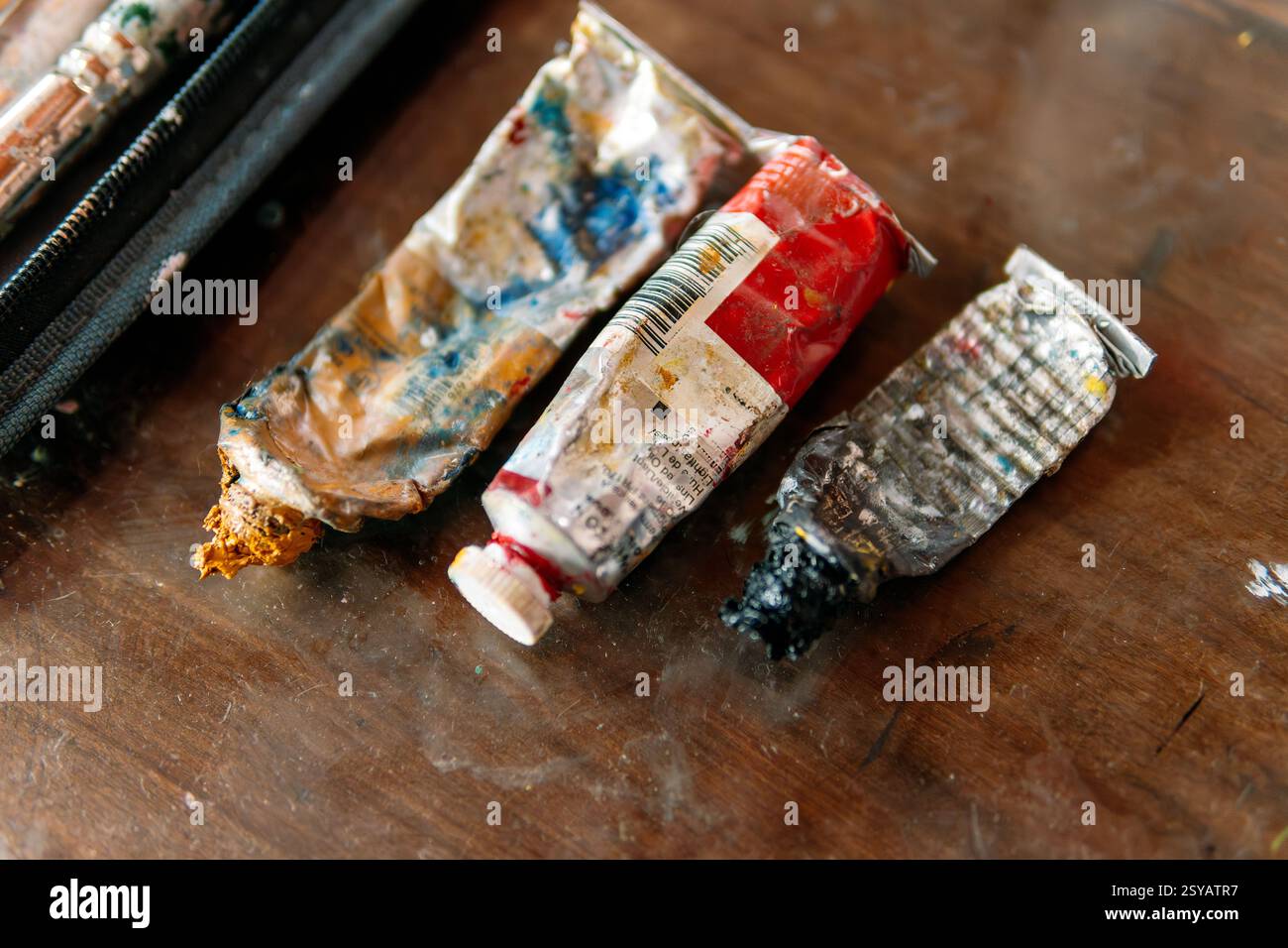 Eine Nahaufnahme verschlissener und zerknitterter Farbröhren, die mit getrockneter Farbe bedeckt sind und auf einem Holztisch liegen. Die unordentlichen Texturen und leuchtenden Farben heben den cre hervor Stockfoto